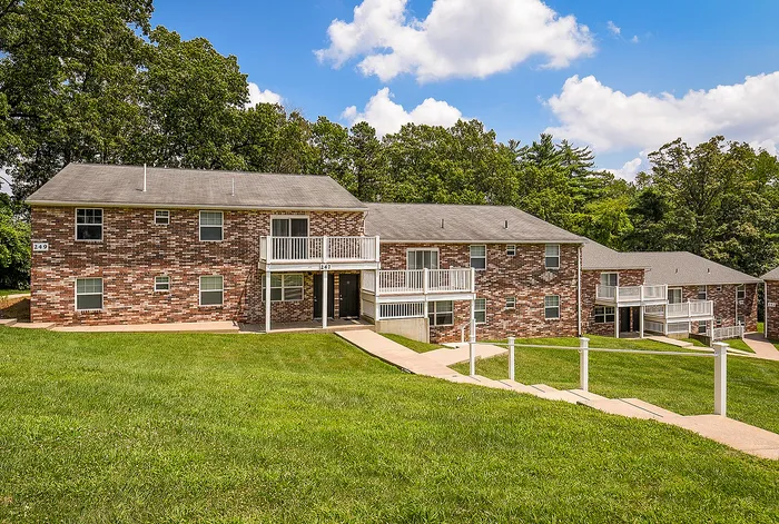Charming brick apartment complex with private balconies, nestled in lush greenery under a vibrant blue sky.