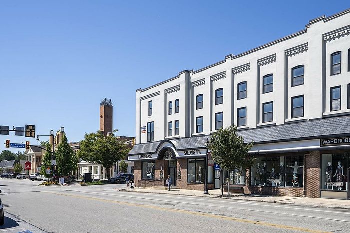 Charming urban street view featuring an elegant white building with retail shops on ground level, clear blue skies, and vibrant trees lining the sidewalk.