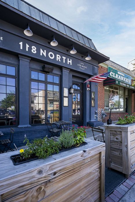 Charming street view of a modern building featuring outdoor seating and vibrant planters, perfect for enjoying food, live music, and drinks.