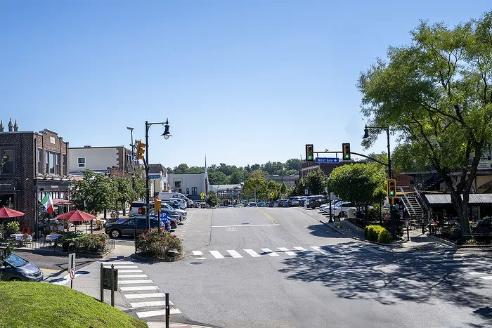 Charming city street view capturing a lively scene with cozy cafes featuring red umbrellas, ample parking, and lush greenery under a bright blue sky.