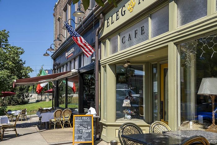 Charming street view of a quaint outdoor cafe with elegant seating, vibrant flags, and inviting storefronts under a clear blue sky.