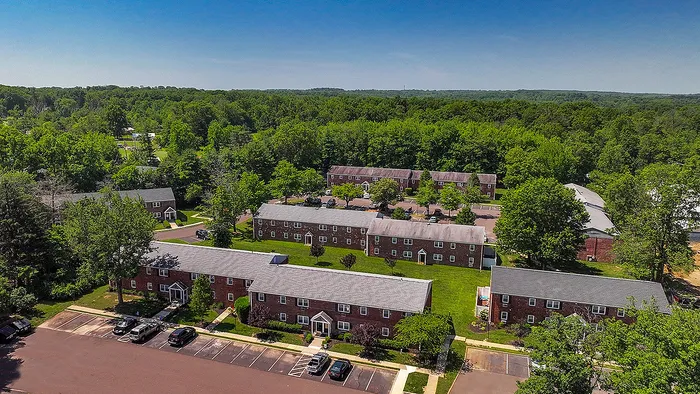 Charming aerial view of a peaceful apartment community surrounded by lush greenery, featuring red brick buildings and ample parking.