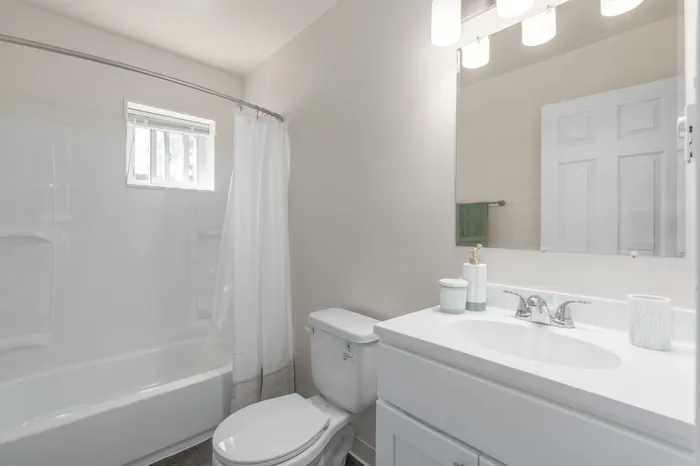 Bright and modern bathroom featuring sleek white fixtures, a large mirror, and a cozy bathtub with natural light.