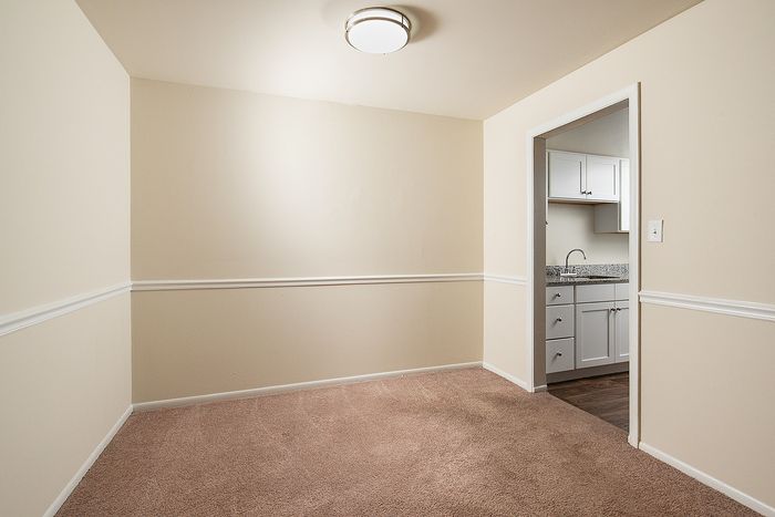 Cozy interior space with soft carpeting, warm neutral walls, modern overhead lighting, and a peek into a stylish kitchen featuring granite countertops and sleek cabinetry.