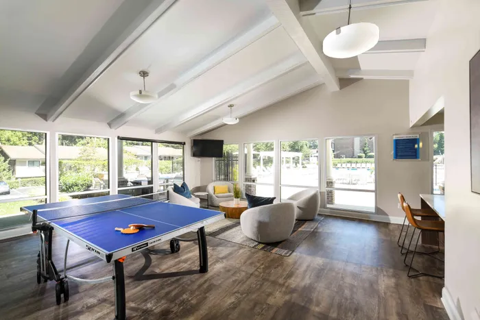 Apartments for Rent in West Chester, PA - Goshen Terrace - Clubhouse with Wood-Style Flooring, Area Rug, Couch, Armchairs, Coffee Table, Side Tables, Ping-Pong Table, Floor-to-Ceiling Windows, and Kitchen Area.