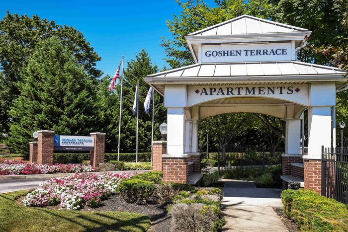 Welcoming entrance to Goshen Terrace Apartments with lush greenery, vibrant flower beds, and prominent signage under a clear blue sky.