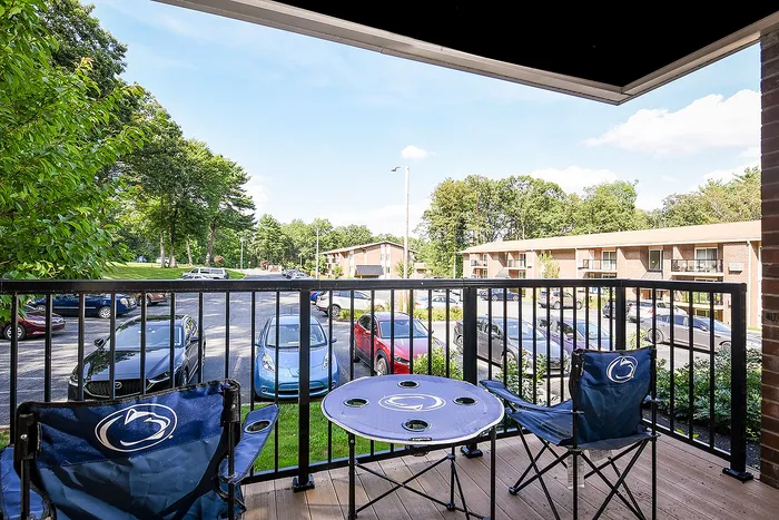 Charming apartment balcony with seating offering scenic views of lush greenery, a serene parking lot, and nearby residential buildings.