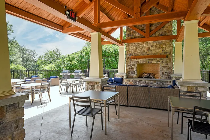 Charming outdoor patio with cozy seating and a rustic stone fireplace, perfect for entertaining. The space includes a sleek wooden pergola and lush green surroundings.