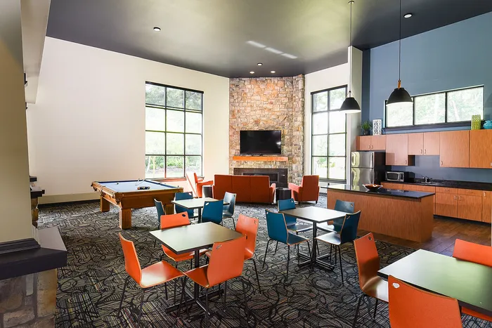 Chic and spacious community lounge featuring modern decor, a pool table, and a cozy stone fireplace. Generous seating area with vibrant chairs and large windows allowing natural light to flood in. Equipped kitchenette perfect for entertaining.