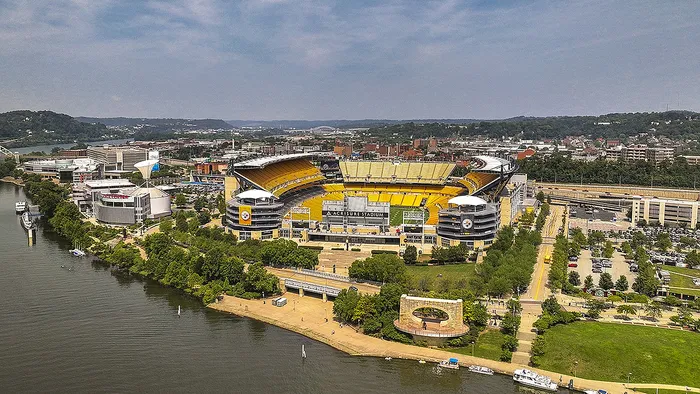 Aerial view of a bustling riverside stadium surrounded by lush greenery and cityscape, showcasing vibrant urban energy.