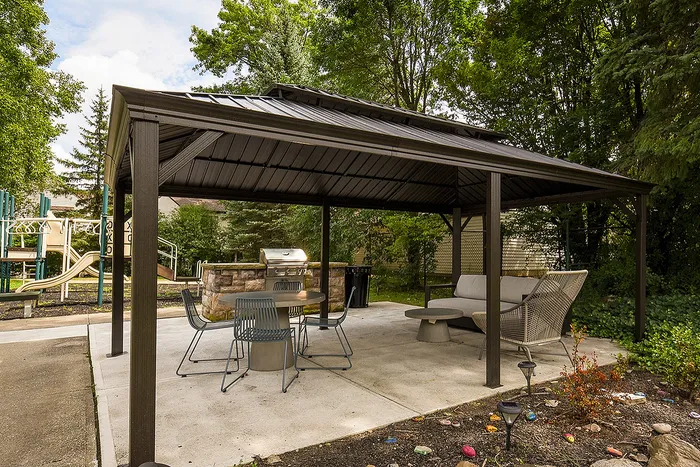 Charming outdoor gazebo with modern seating and BBQ grill, nestled in lush greenery near a playground.