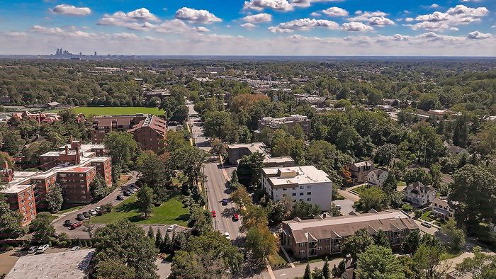 Breathtaking aerial view of a vibrant neighborhood surrounded by lush greenery, showcasing mid-rise apartment buildings and a distant city skyline under a blue sky with fluffy clouds.