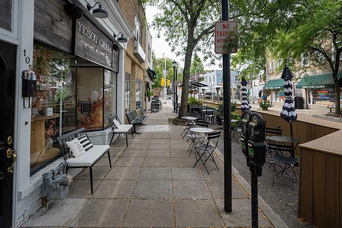 Charming street view featuring a cozy Greek market with outdoor seating, vibrant greenery, and inviting sidewalk ambiance.