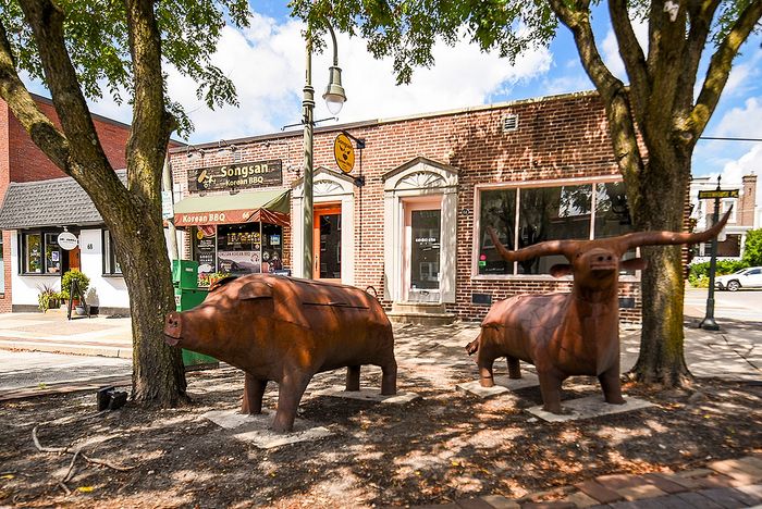Charming street scene featuring a Korean BBQ restaurant with metal animal sculptures under lush trees, capturing a blend of culture and art.