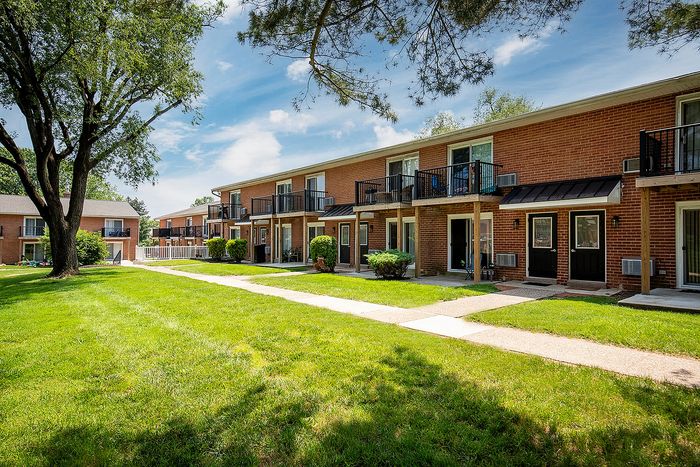Charming brick apartment complex with private balconies set amidst a lush green lawn and mature trees, perfect for serene living.