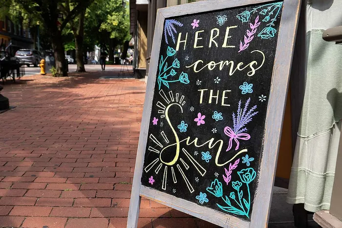 Charming sidewalk chalkboard sign with artistic floral designs, displaying the cheerful message 'Here Comes the Sun' on a sunny, tree-lined street.