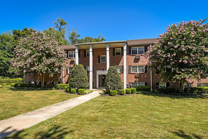 Charming brick apartment building with lush landscaping and blooming trees against a clear blue sky, offering a serene and welcoming atmosphere.