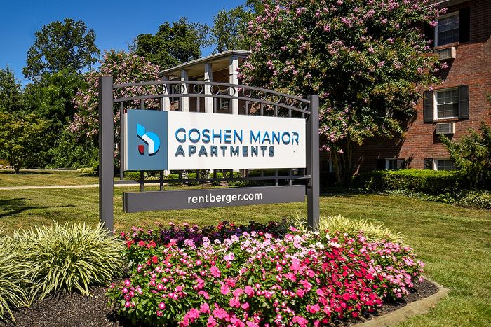 Charming sign for Goshen Manor Apartments surrounded by vibrant flower beds and lush greenery, promising a welcoming living environment.