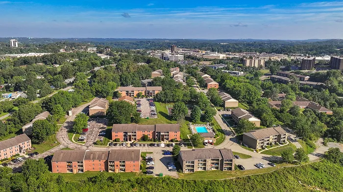 Vibrant aerial view of a lush apartment community with spacious layouts, ample parking, and a refreshing swimming pool, nestled in a picturesque landscape.
