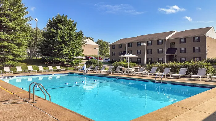 Inviting outdoor pool area with sun loungers, nestled among lush greenery beside a charming apartment building under a clear blue sky.