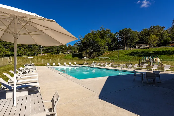 One-Bedroom Apartments in Bethel Park, PA - Lindenbrooke - Swimming Pool with Expansive Sundeck, Lounge Chairs, Tables and Chairs with Umbrellas, and Surrounding Manicured Landscape.