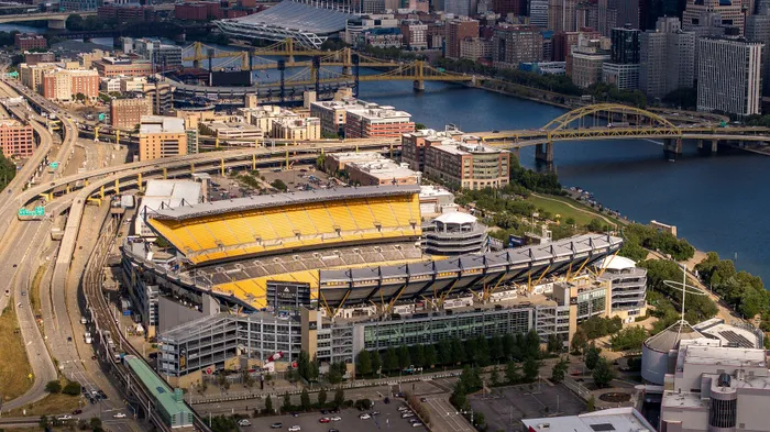 Aerial view of a vibrant cityscape featuring a prominent stadium with striking yellow seats, surrounded by bustling roads and graceful bridges over a scenic river.
