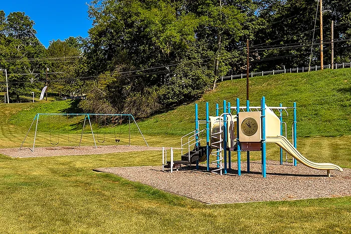 V Dog-Friendly Apartments in Bethel Park, PA - Lindenbrooke - Playground with Wood Chips-Covered Surface, Jungle Gym with Slide, Swing Set, and Surrounding Manicured Landscape.