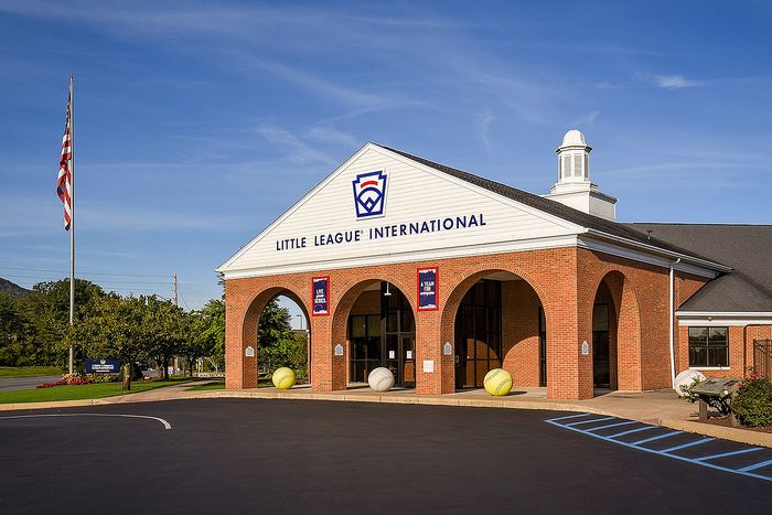 Elegant brick building with arched entryways, displaying the Little League International sign, surrounded by a vibrant outdoor landscape with flag in the foreground.
