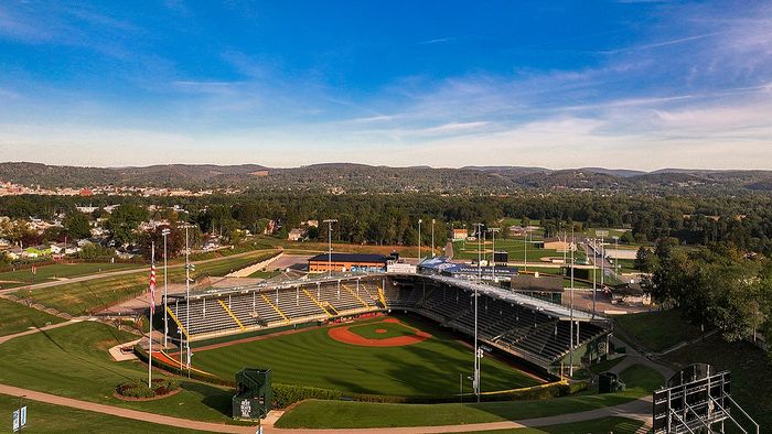 Aerial view of a picturesque baseball stadium surrounded by lush greenery and scenic hills under a clear blue sky.