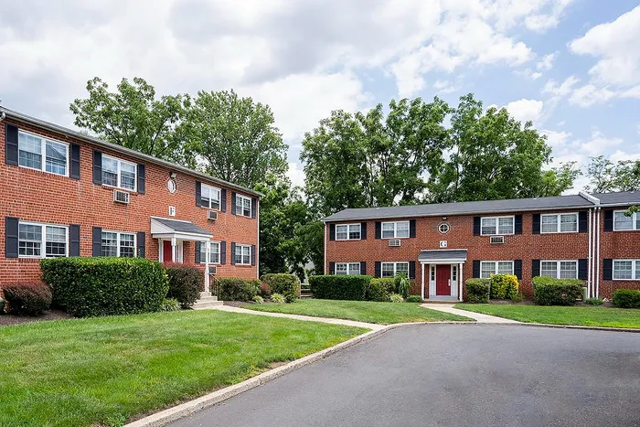 Charming brick apartment buildings with lush green landscaping and tree-lined pathways under a bright blue sky.