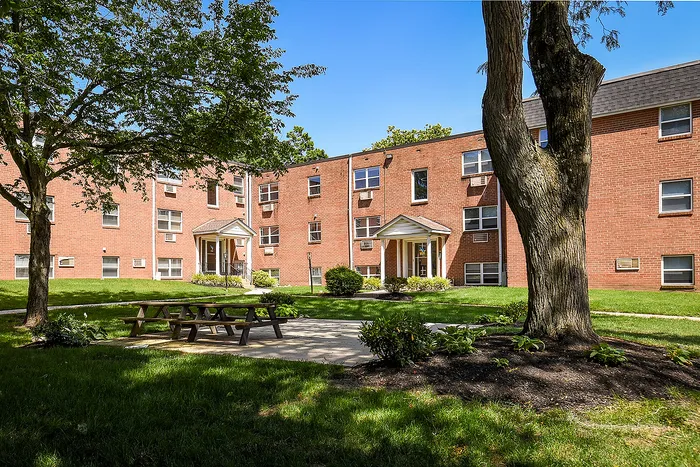 Charming brick apartment building with lush green courtyard, featuring mature trees and a welcoming picnic area.