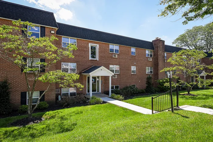 Charming brick apartment building with lush landscaped grounds and inviting white entryway under a bright blue sky.