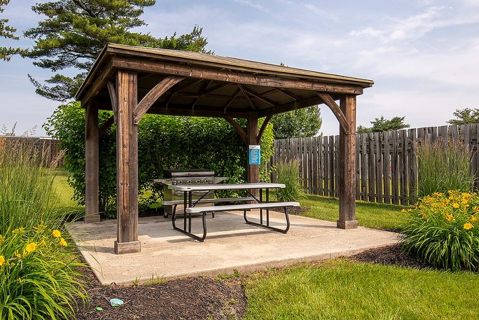 Charming outdoor pavilion with a rustic wooden structure, picnic table, and BBQ grill set in a lush garden with flowering plants and a wooden fence backdrop.