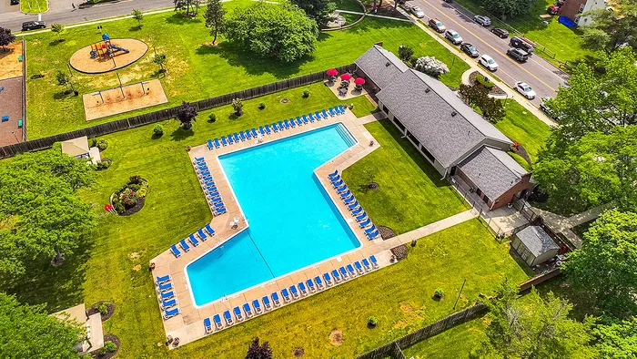 Vibrant aerial view of a luxurious apartment complex featuring a large, inviting swimming pool surrounded by blue lounge chairs, lush green lawns, and a charming playground area for family-friendly enjoyment.