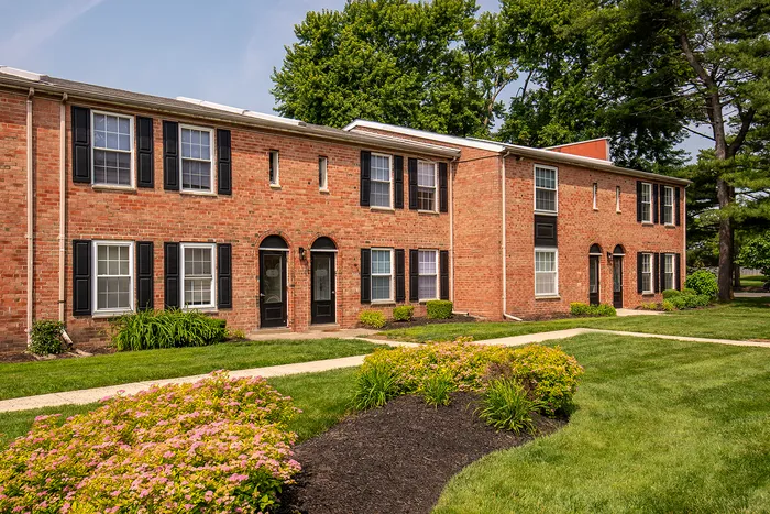 Charming red brick apartment complex surrounded by lush greenery and vibrant gardens, featuring classic black shutters and elegant archway entrances.