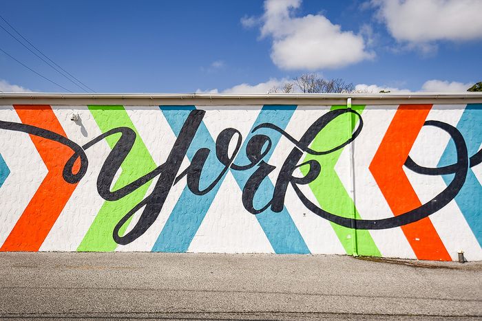 Vibrant street mural with flowing script and bold diagonal stripes in orange, green, and blue, under a clear blue sky.