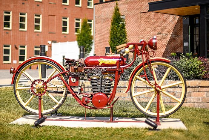 Vibrant vintage red motorcycle sculpture on display against a charming brick building backdrop, showcasing intricate design and historic appeal.