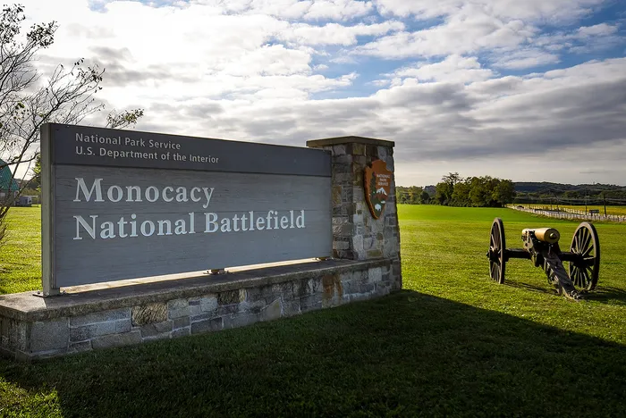 Scenic view of Monocacy National Battlefield sign with historic cannon and expansive green landscape under a partly cloudy sky.