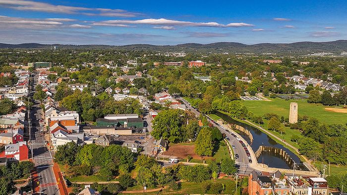 Charming aerial view of a vibrant community with tree-lined streets, lush greenery, and a historic tower by a peaceful canal, set against rolling hills and a bright blue sky.