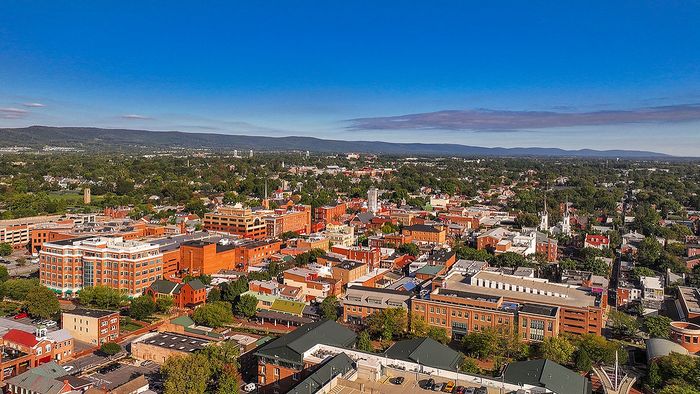 Aerial view of a vibrant town with picturesque buildings, lush greenery, and scenic mountains in the background under a clear blue sky.