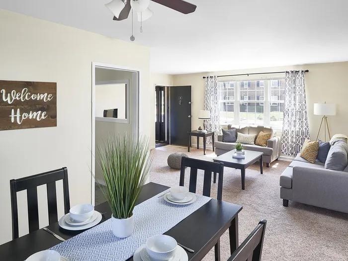 Bright and welcoming living room with modern decor, cozy gray sofas, and a stylish dining area with a charming 'Welcome Home' sign.