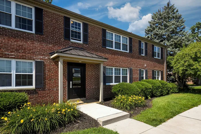 Charming two-story brick apartment building with elegant black shutters and a welcoming front entrance, surrounded by lush greenery and vibrant yellow flowers.