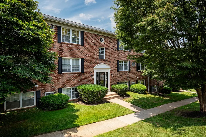 Charming brick apartment building surrounded by lush greenery and a well-maintained lawn, perfect for comfortable living.
