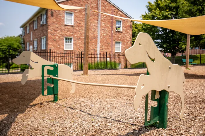 Inviting community playground with charming dog-themed seesaw, surrounded by lush greenery and a brick building backdrop.
