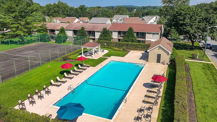 Inviting community view showcasing a large swimming pool with lounge chairs and vibrant umbrellas, tennis courts, and charming homes surrounded by lush greenery.