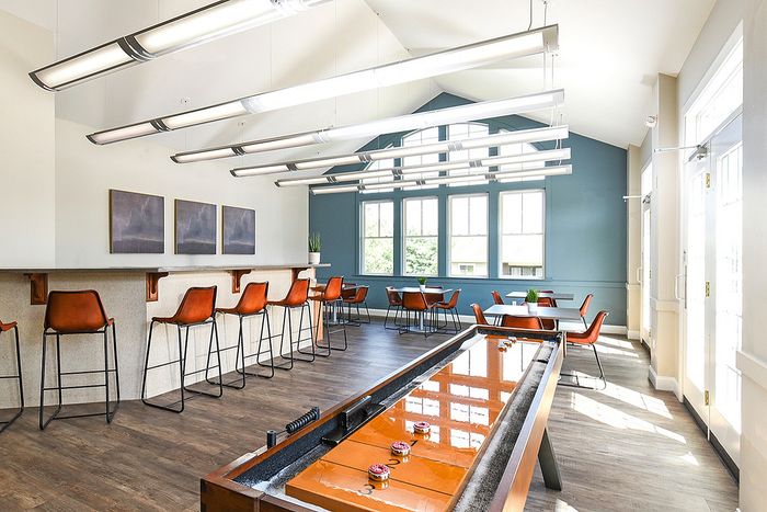 Modern, sunlit community lounge featuring high ceilings, sleek bar seating, and stylish decor with a shuffleboard table.