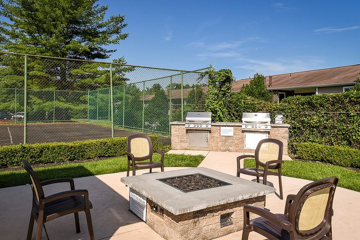 Inviting outdoor patio featuring a modern fire pit, comfortable seating, and dual grills, nestled beside a tennis court under a clear blue sky.
