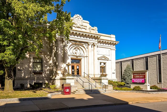 Elegant historic library with grand stone architecture, flanked by lush green trees and a welcoming entrance, under a clear blue sky.