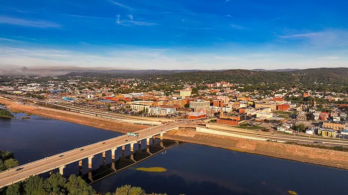 Breathtaking aerial view of a vibrant cityscape with a stunning bridge over a serene river, framed by lush green hills and a brilliant blue sky.