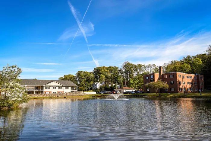 Charming lakeside apartment complex with fountain, surrounded by lush trees and clear blue skies.